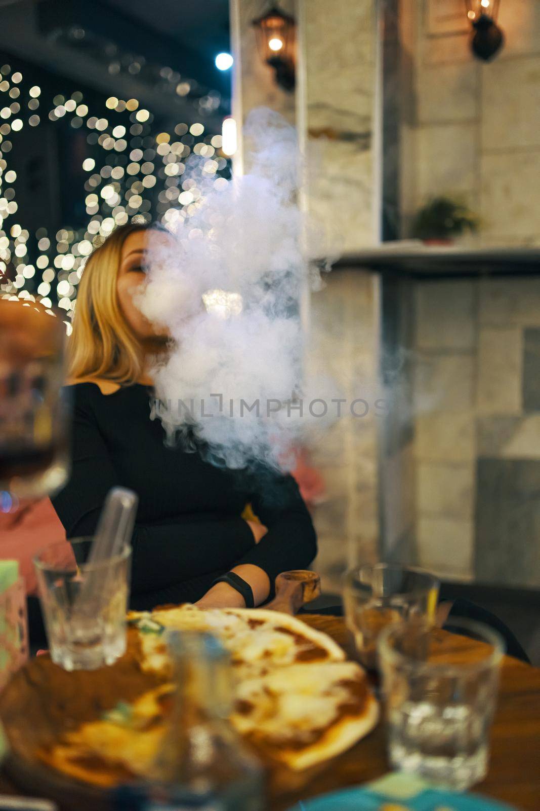 Beautiful woman smoking hookah in pub. Woman in black dress is enjoying great evening in cafe. Party at lounge bar. Pizza and glasses on table.