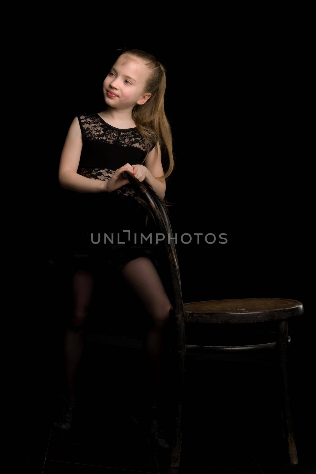 Portrait of a little girl who stands near an old Vienna chair on a black background. Studio photo on the cover of the magazine.