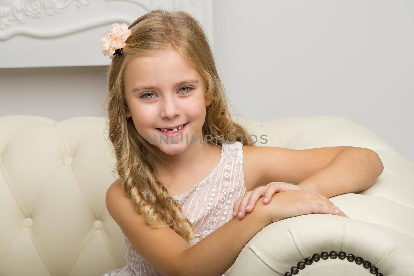 Beautiful happy girl sitting on sofa. Retro style closeup portrait of smiling blonde girl in nice dress sitting on comfortable couch. Adorable happy seven years old kid posing in studio