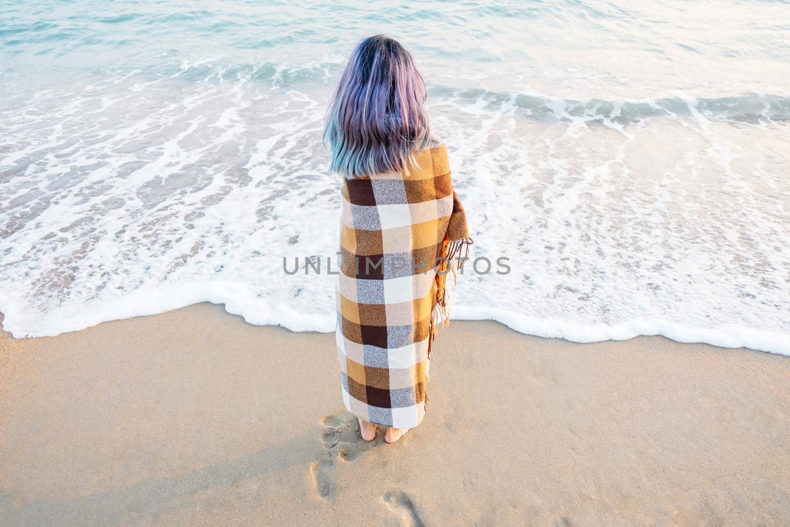 Summer vacations. Young woman wrapped in a plaid standing on sand shore near the sea.