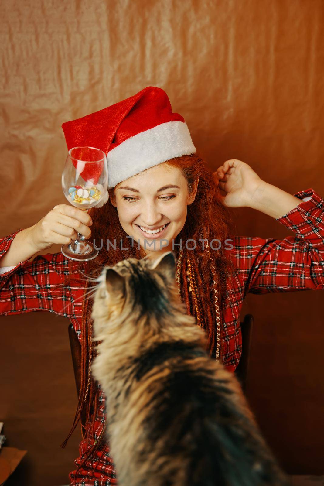 Smiling girl in santa hat hold glass of tablets and play with fluffy cat. Red-haired female with dreads in plaid pajamas. Christmas in quarantine.