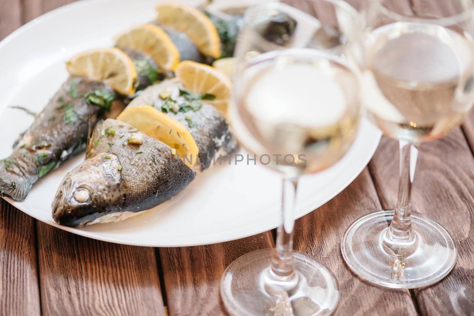 Simple fish dish with lemons and greenery on a white plate on wooden table with two glasses of white wine.
