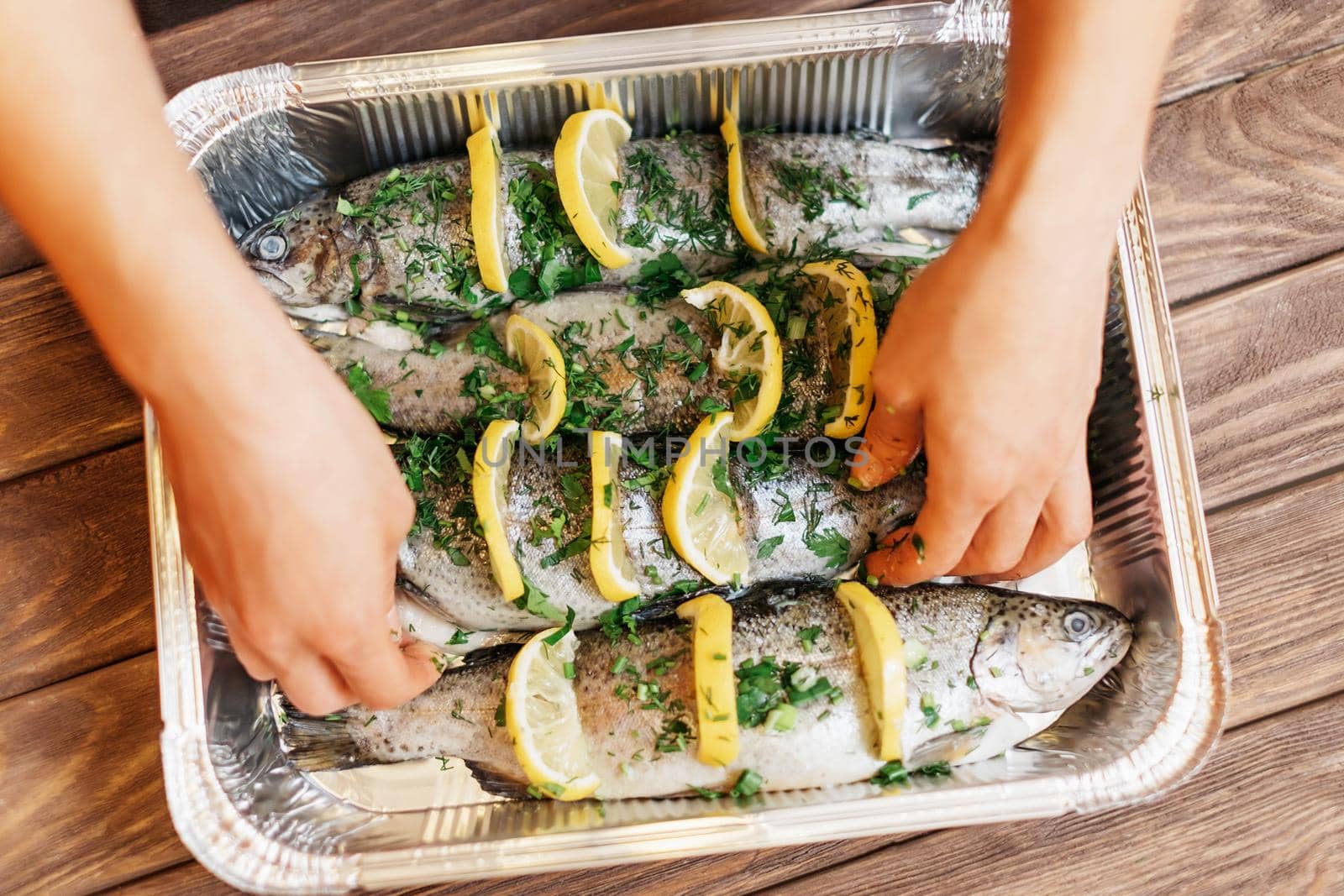 Unrecognizable woman preparing fish trout with lemons and greenery on foil tray, view of hands.