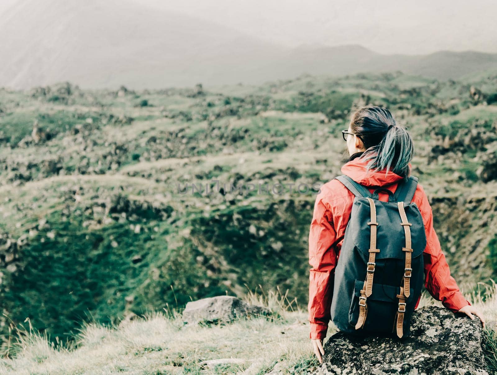 Explorer backpacker young woman resting on rocky stone outdoor, rear view.