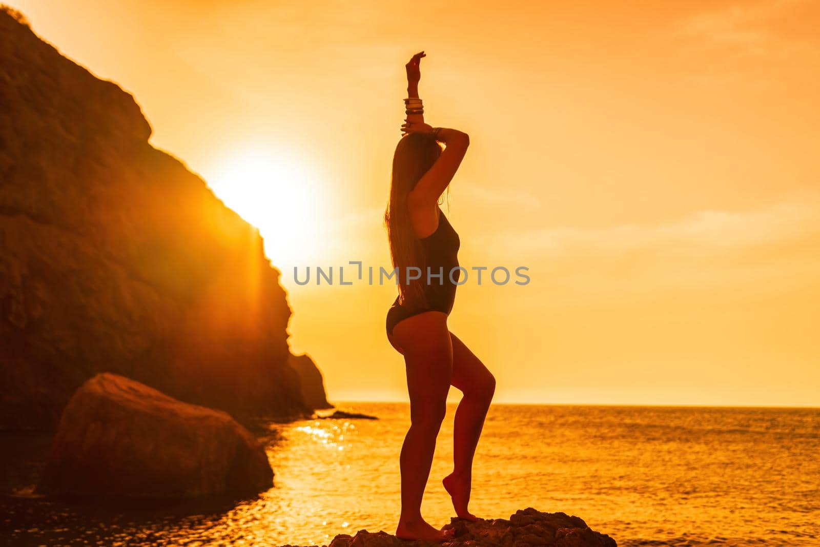 Young woman with long hair in black swimsuit and boho style braclets practicing outdoors on yoga mat by the sea on a sunset. Women's yoga fitness routine. Healthy lifestyle, harmony and meditation by panophotograph