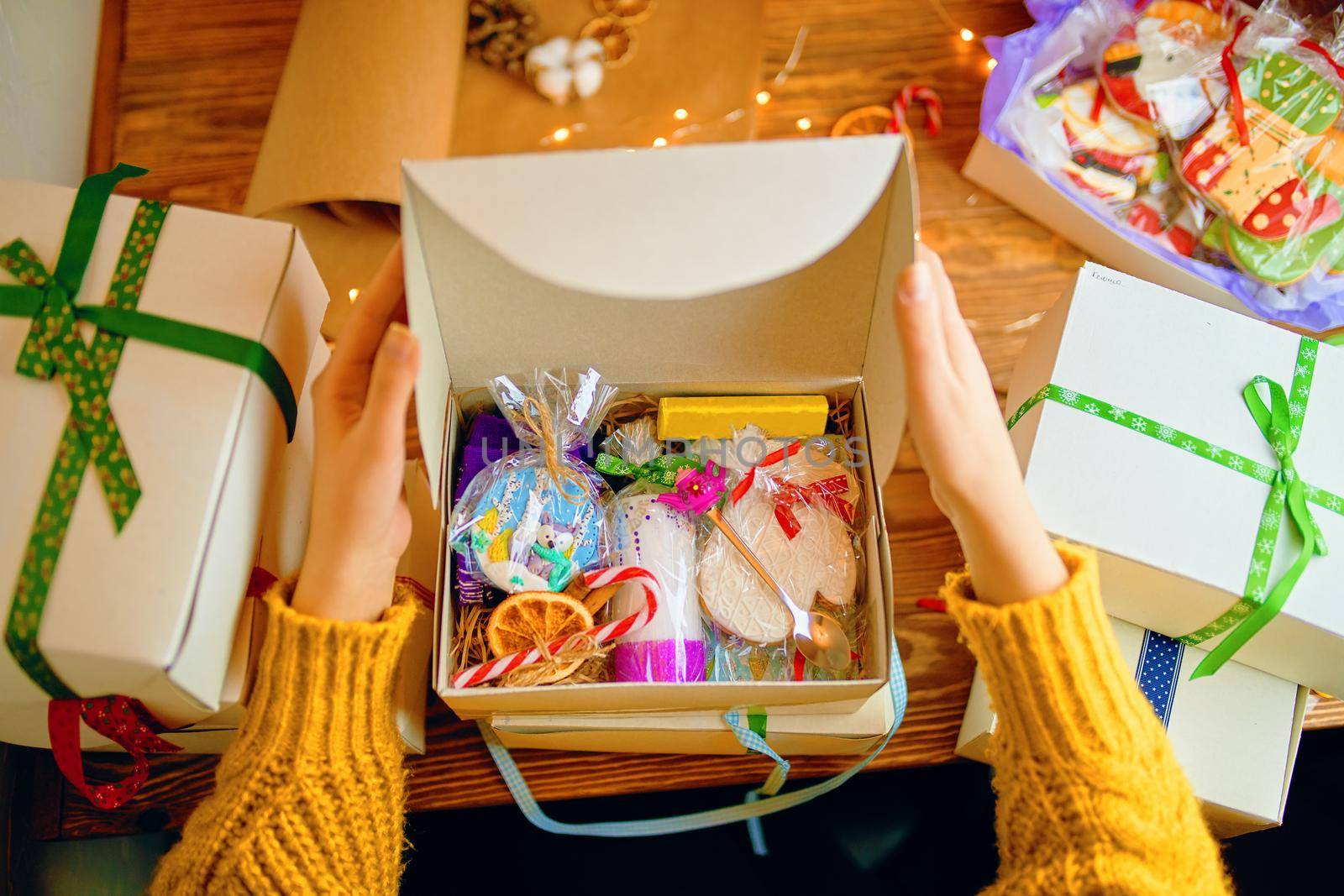 Women's hands in knitted sweater open Christmas gift. Gingerbread with icing, candle with ornamental, souvenir made of polymer clay and decorations in box. Boxes on wooden table.