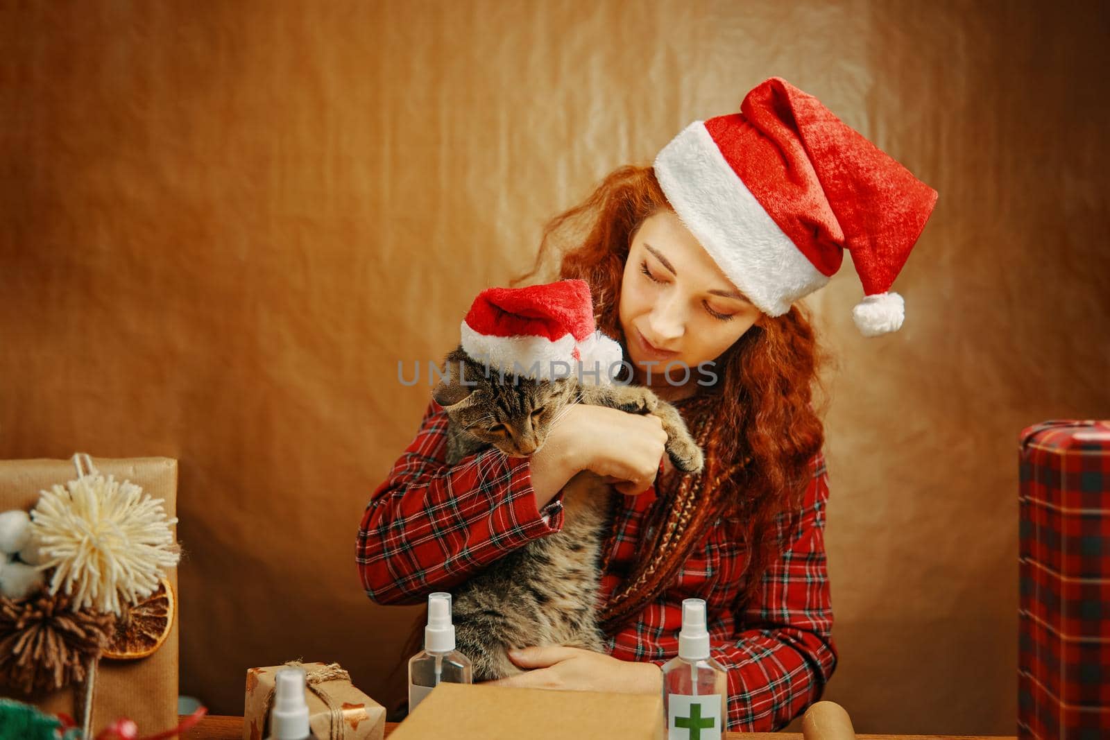 Beautiful girl in Christmas hat with cat in Santa Claus hat near table with gift boxes and sanitizers. Red-haired woman with dreadlocks in plaid pajamas. New year's atmosphere. Festive accessories.