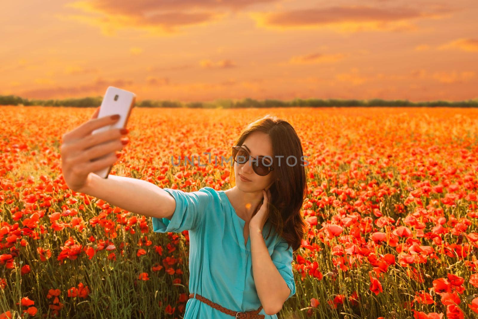 Beautiful young woman taking selfie with smartphone on background of red poppies meadow in summer at sunset.