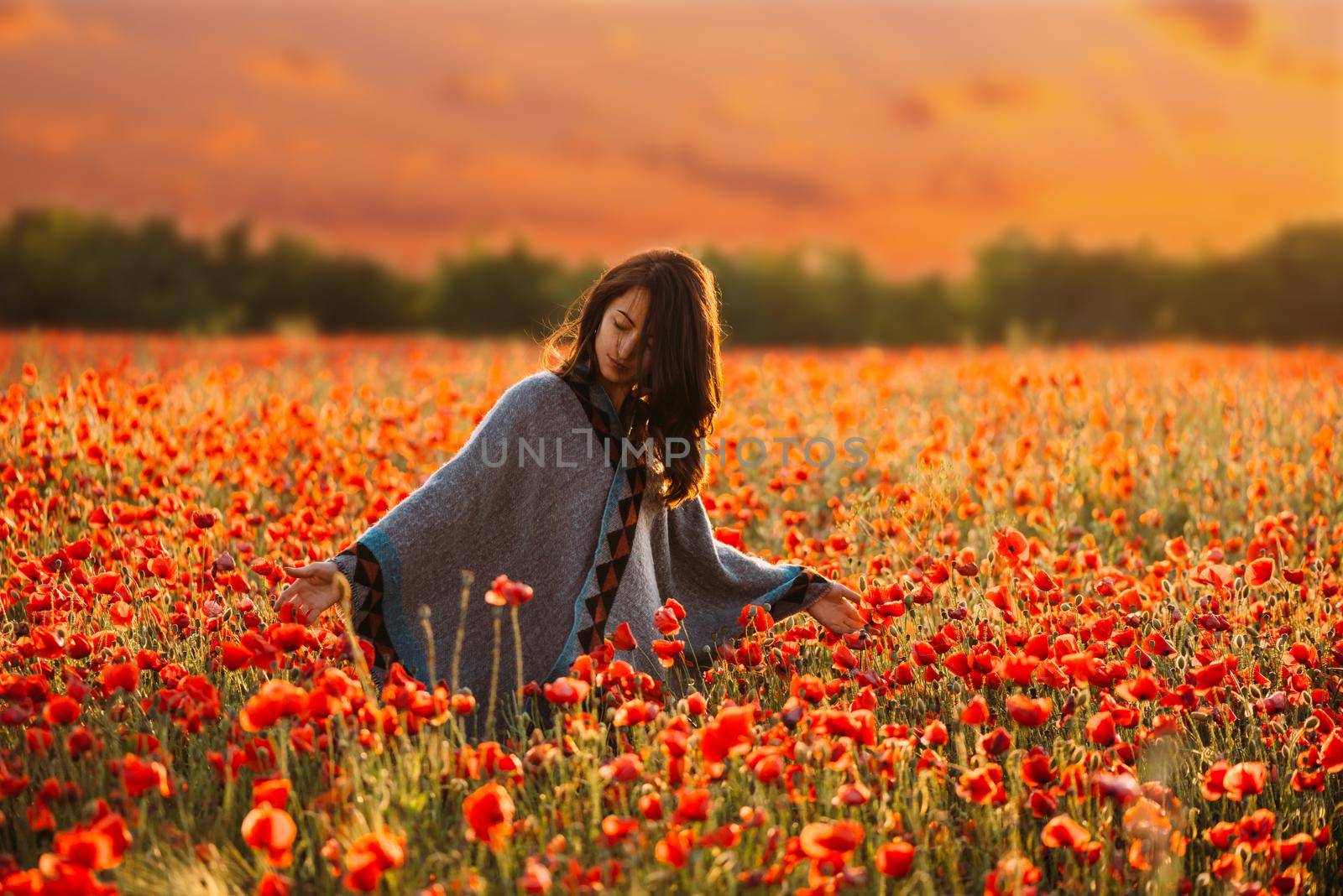 Beautiful young woman walking in flower meadow and touching red poppies at sunset in summer.
