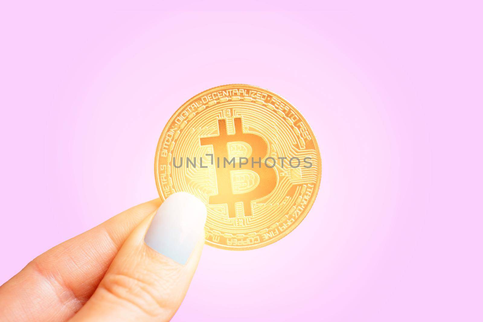 Female hand holding glowing gold bitcoin on a pink background, symbol of virtual money.
