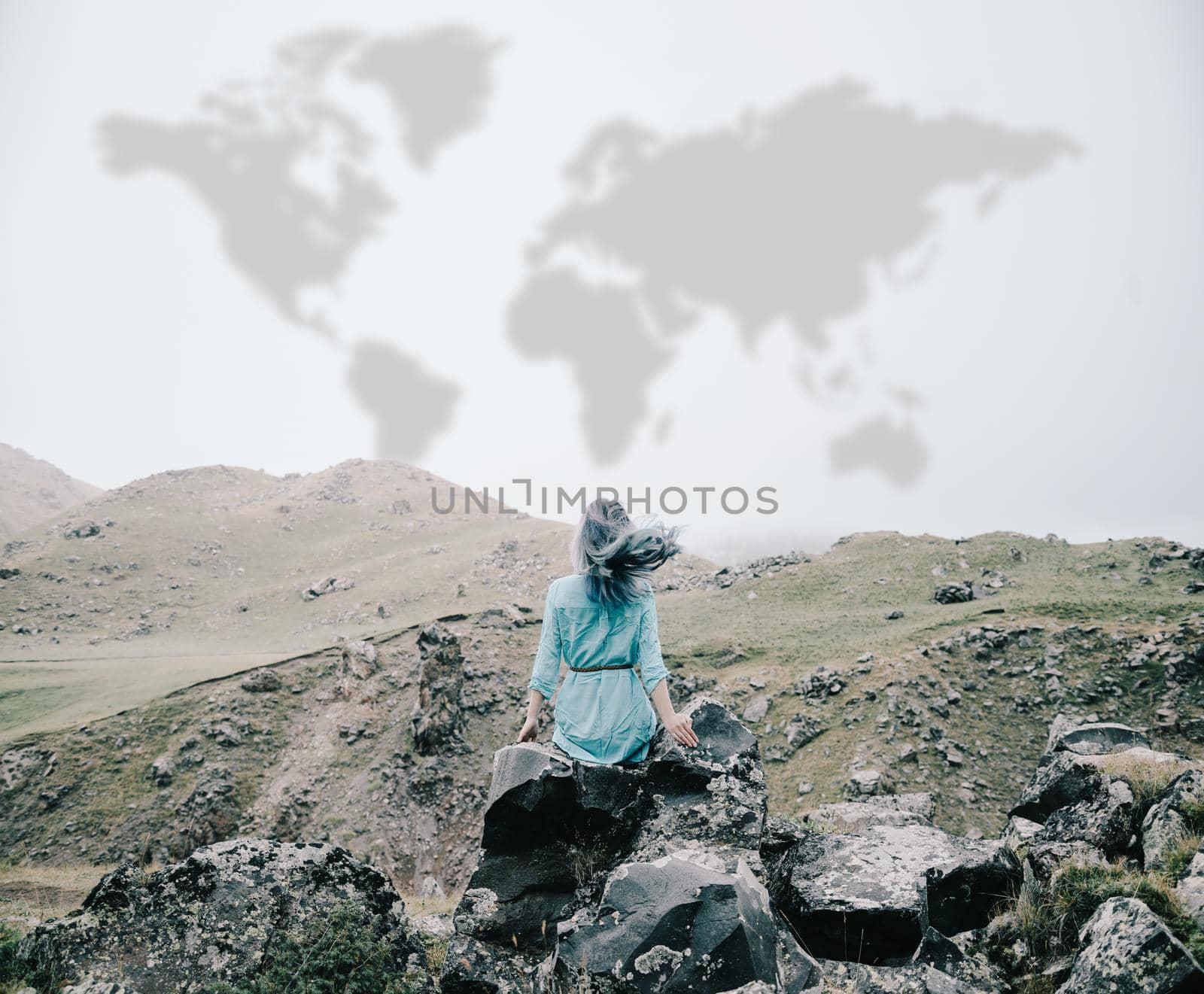 Young woman sitting on rocky mountain outdoor, rear view. Map of world is painted on sky. Concept of travel.