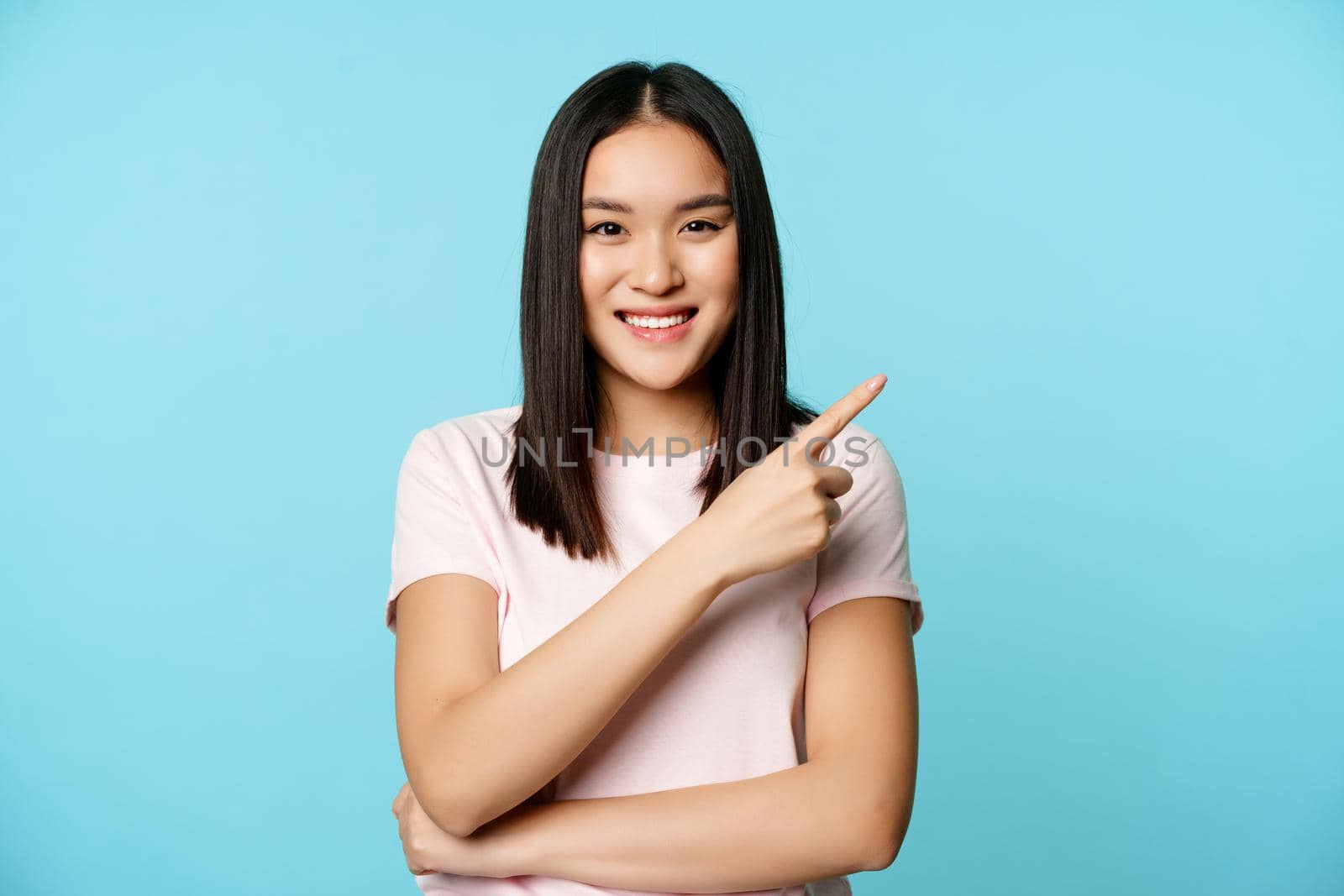 Smiling young asian woman 20 years old, pointing finger at upper right corner, showing promo banner, blue background.