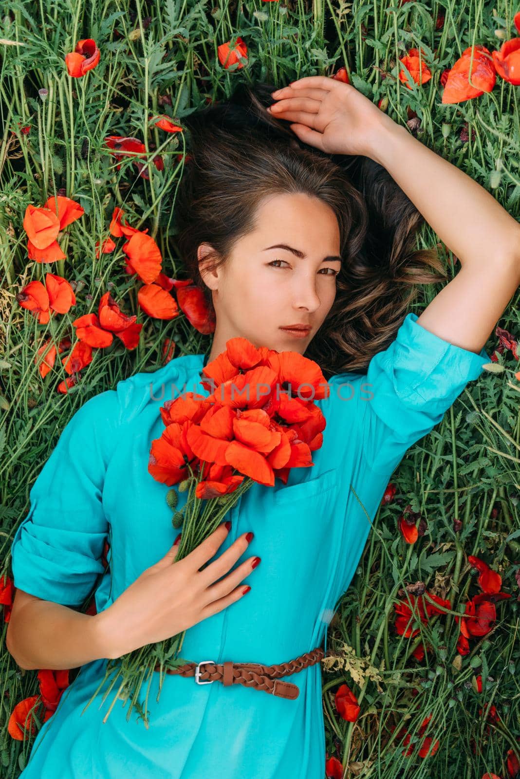 Attractive young woman lying on red poppy meadow with bouquet of flowers, looking at camera. Beauty and fashion concept.