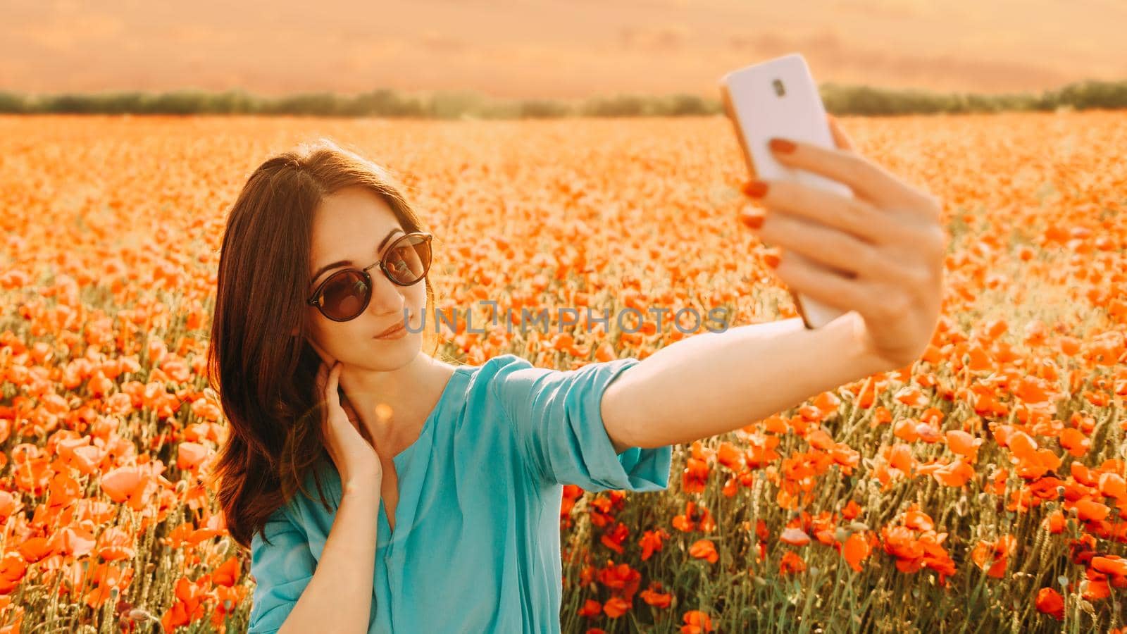 Beautiful girl making selfie with smartphone on background of poppies flower meadow in summer at sunset.