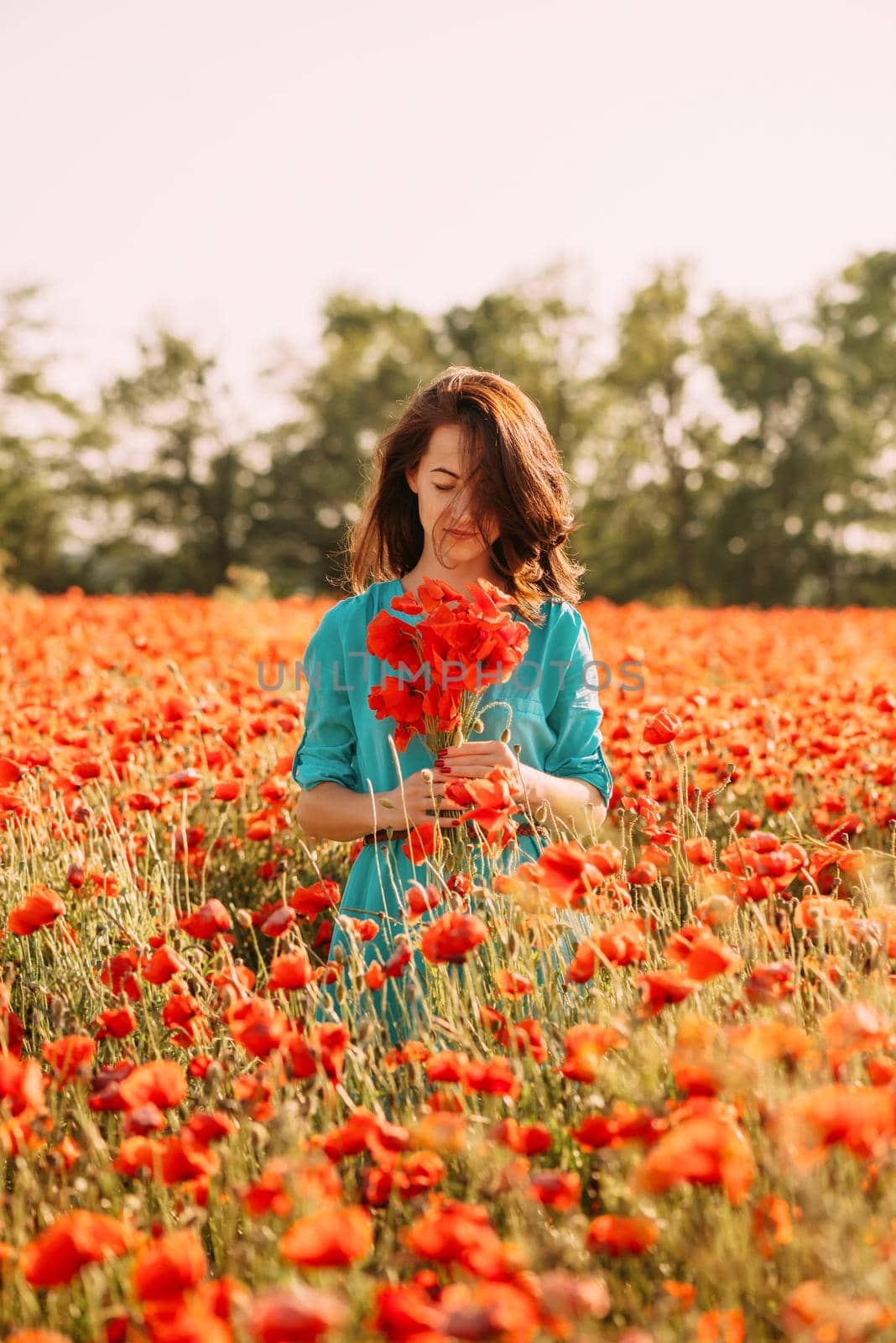 Beautiful brunette young woman standing in red poppies field with bouquet of flowers on sunny summer day.
