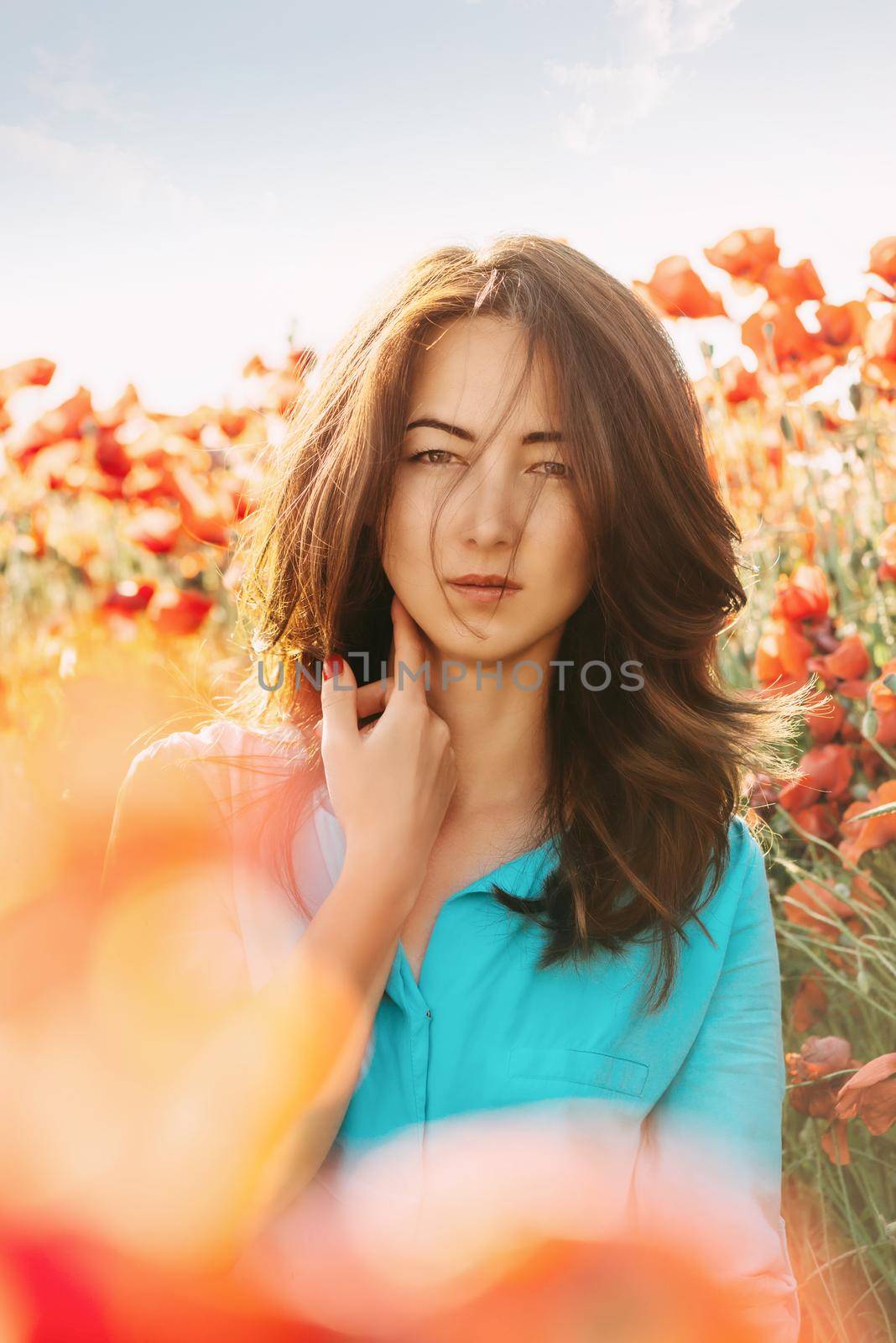 Portrait of beautiful brunette young woman in red poppies meadow, looking at camera.