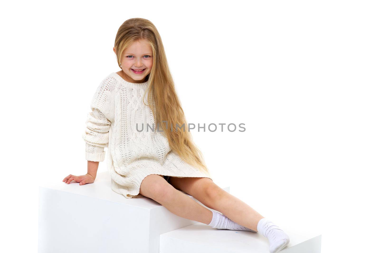 Cute little girl wearing warm knitted dress sitting on white staircase. Happy smiling long haired girl posing in studio against white background. Happy childhood concept