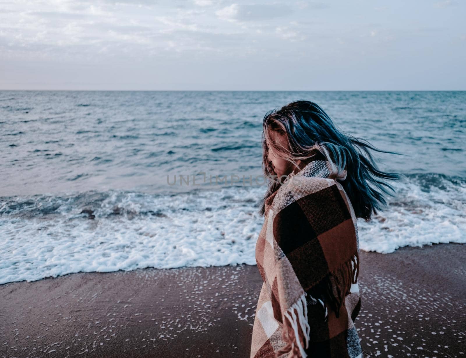 Young woman wrapped in plaid walking on sand coast near the sea in windy weather.