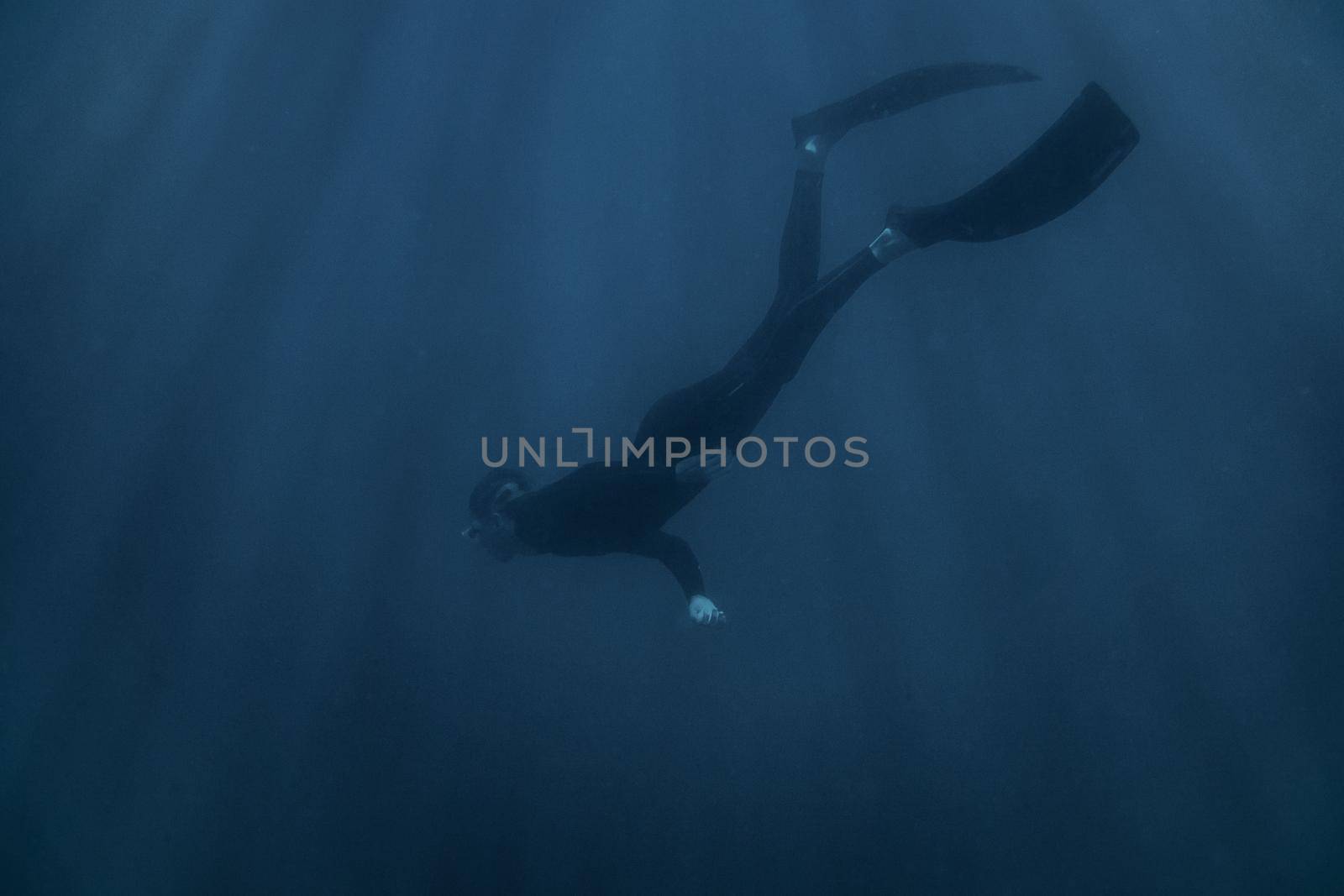 Young man freediver wearing in wetsuit and flippers swimming underwater in blue deep sea.