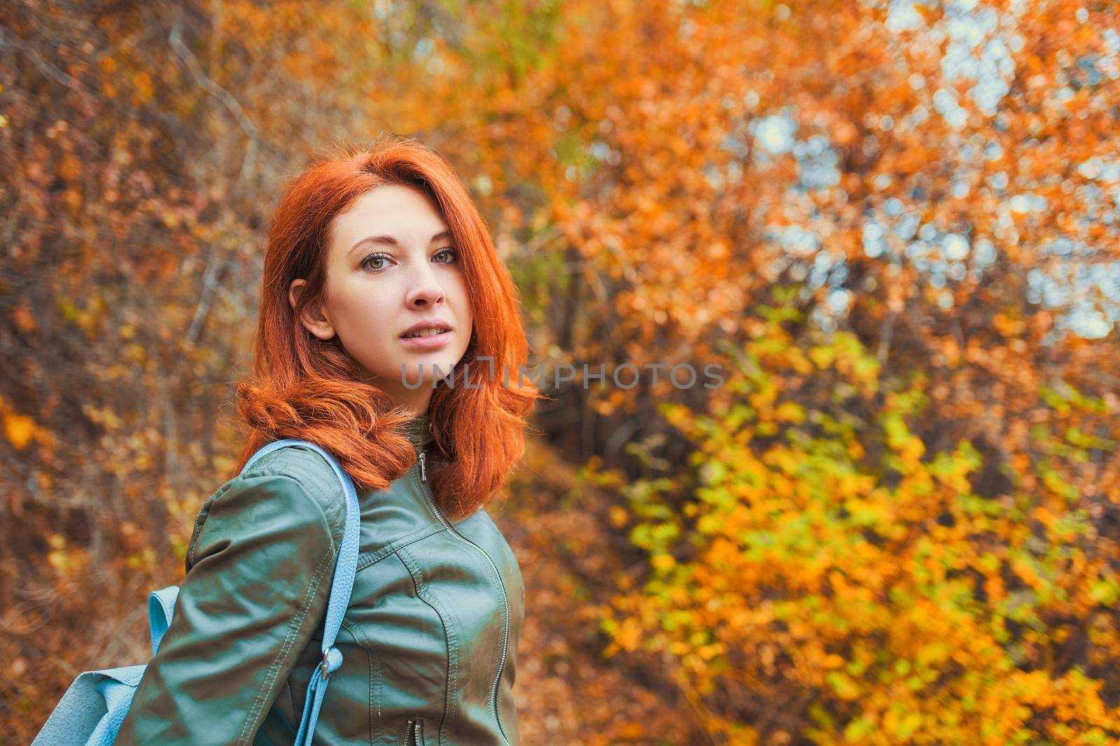 Red-haired woman on the background of the autumn forest. Amazing woman on vacation. Mountain fall forest. A trip to nature. Leather jacket and fashionable backpack.