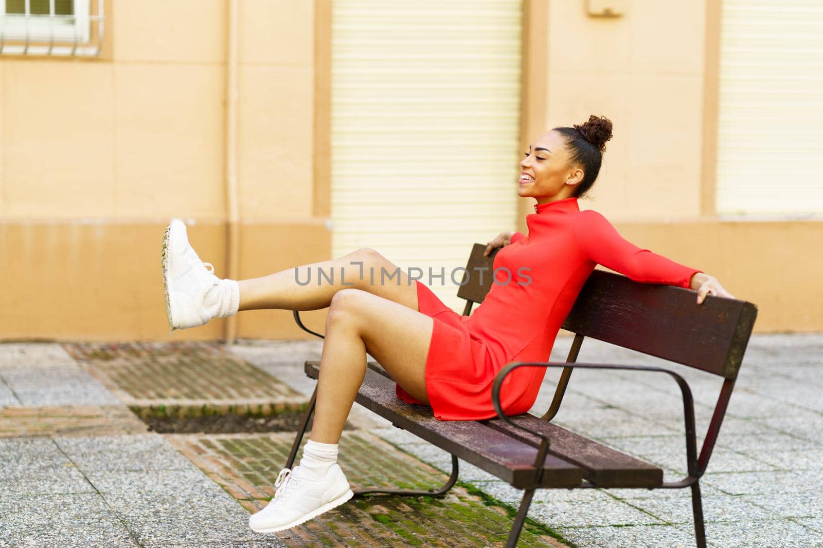 Happy mixed woman, moving her legs in joy, sitting on a bench in the street. by javiindy