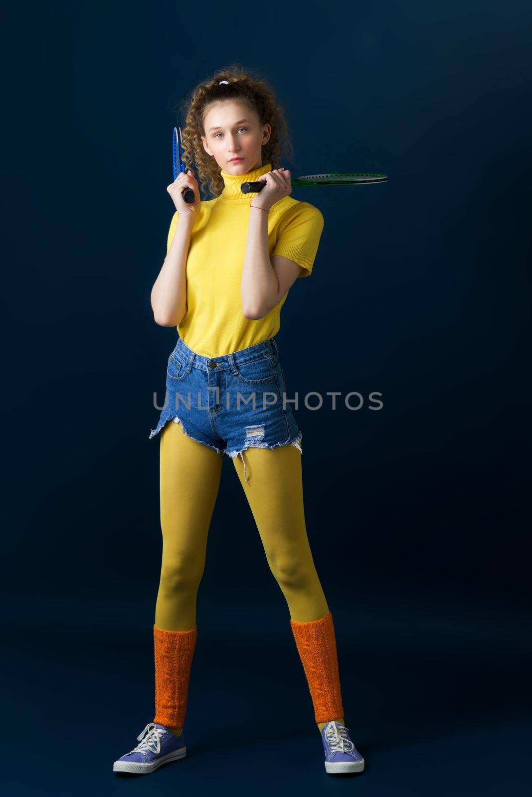 Shot of a beautiful tennis player holding a racket. Happy smiling girl doing sports in a bright yellow turtleneck, leggings, shorts and socks, leggings posing on a blue background