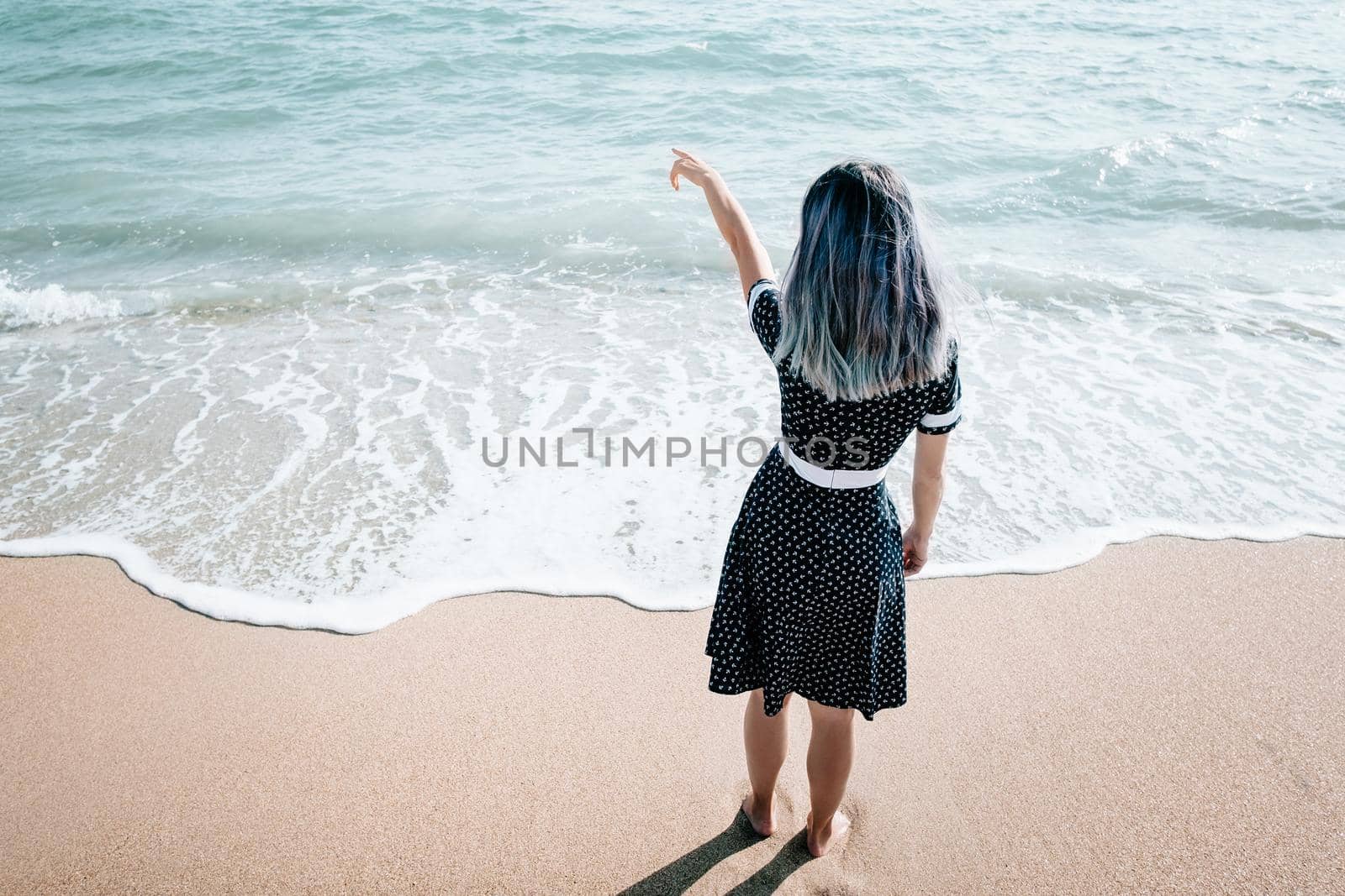 Full length image of young woman in dress standing on sand coast and pointing at sea, beach vacations.