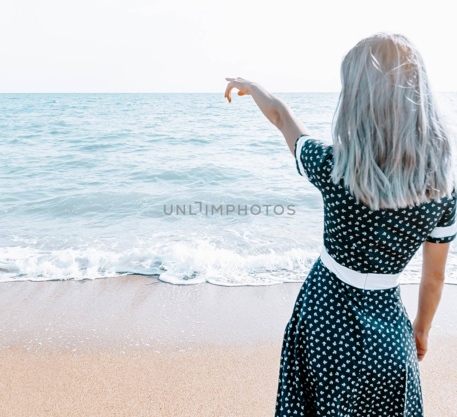 Unrecognizable young woman in dress standing on sand coast and pointing at sea, copy-space.