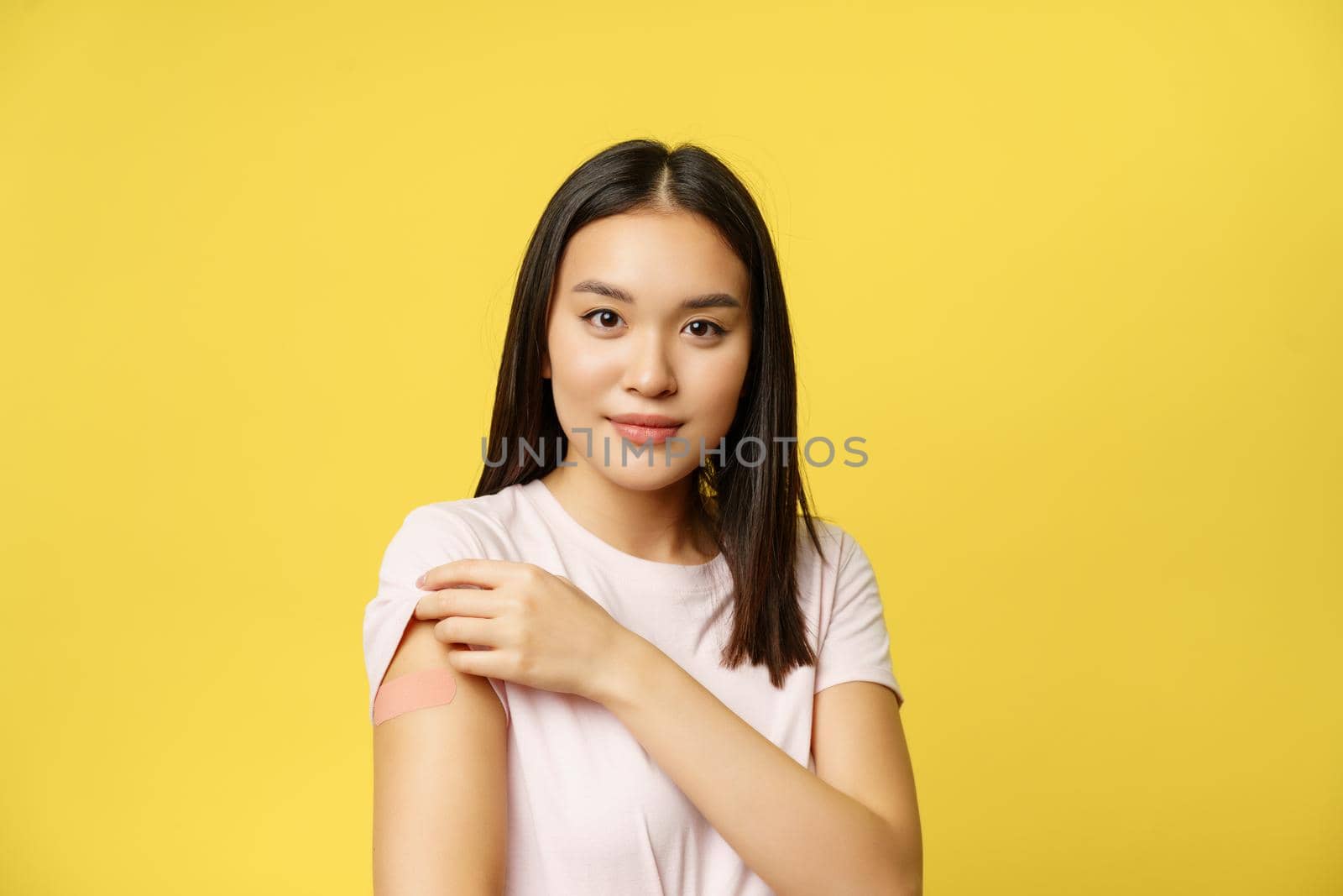 Covid-19 and healthcare medical concept. Smiling korean girl shows shoulder with patch, took shot of coronavirus vaccine during pandemic, yellow background.