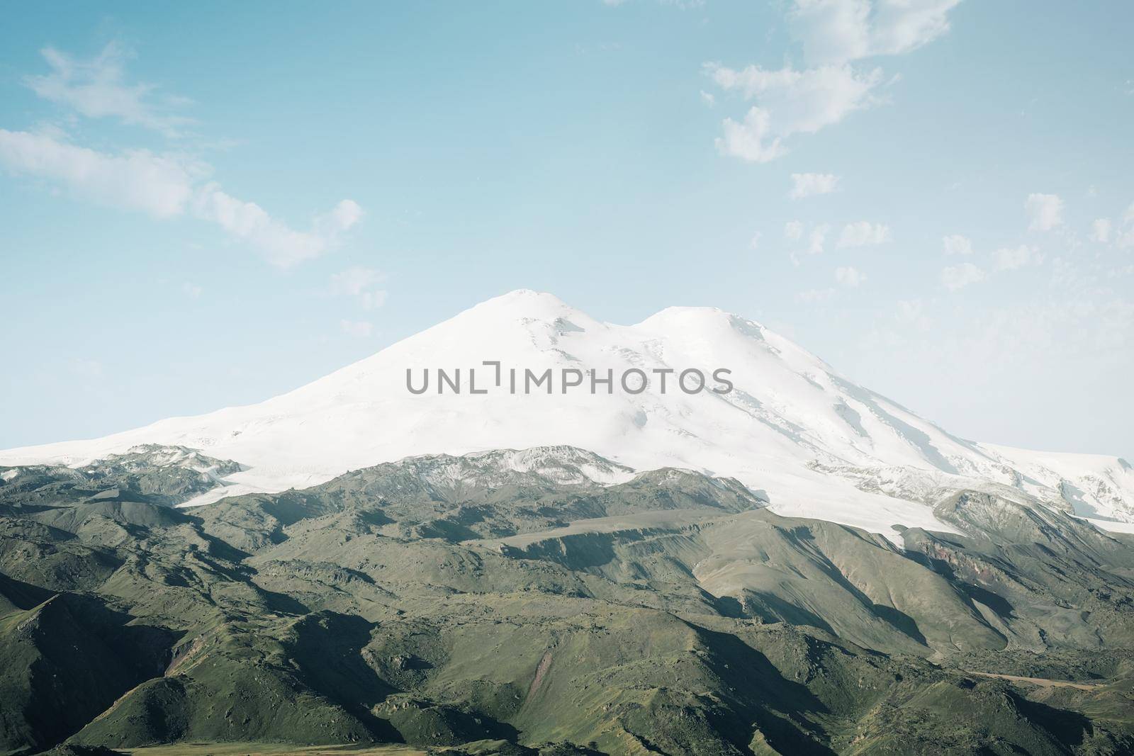 Mount Elbrus with snowy peaks and green fields on sunny day, Caucasus mountain range, Russia.