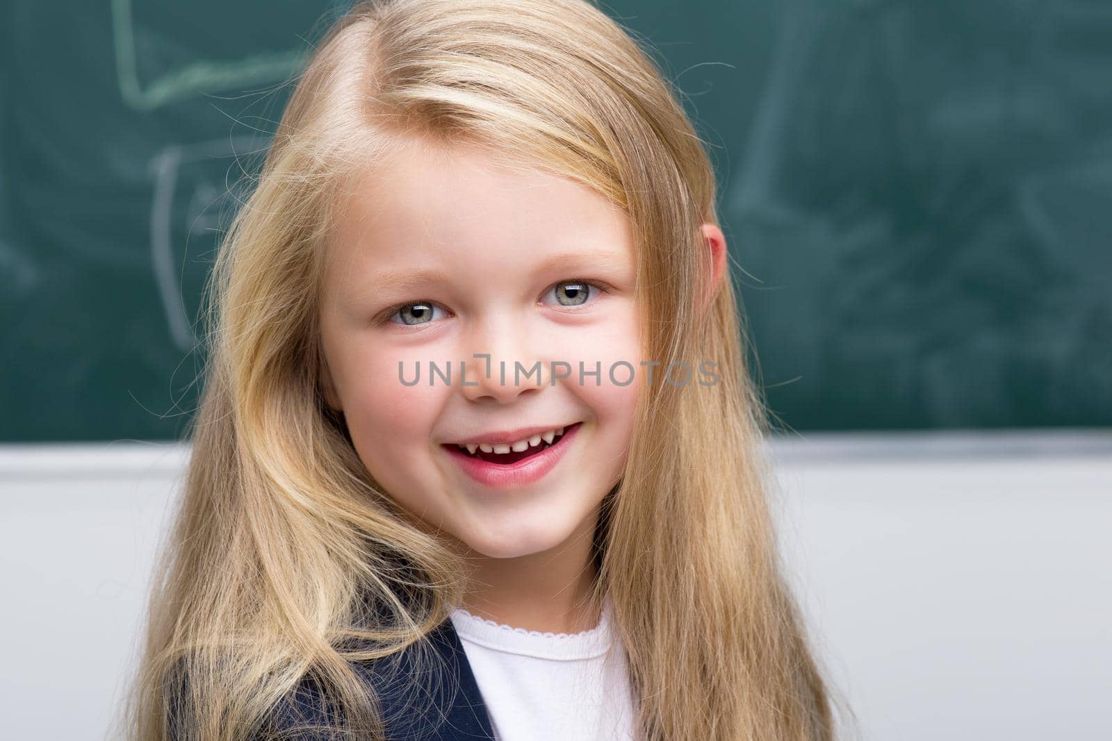 Close up portrait of beautiful girl. Adorable blonde long haired girl posing on background of blackboard in classroom. Cute smiling elementary school student girl. Back to school, education concept