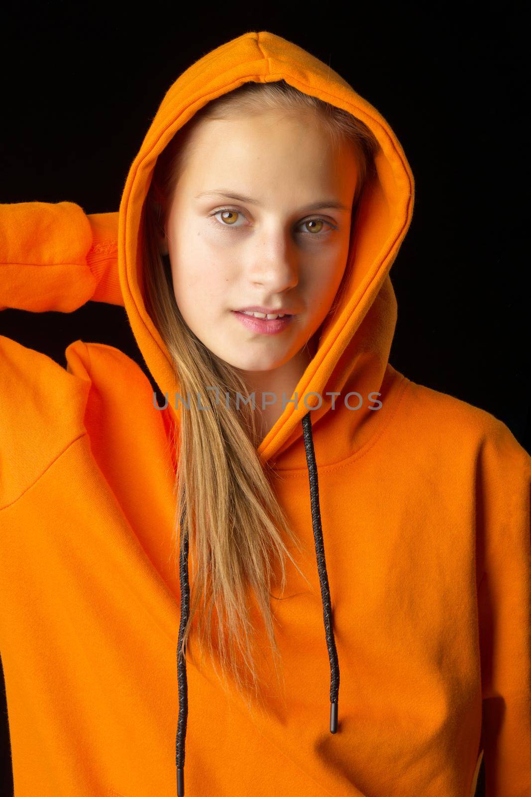 Closeup portrait of cheerful teenage girl in hood. Cheerful teenager wearing orange hoodie posing on black background. Beautiful brown eyed girl staring at camera