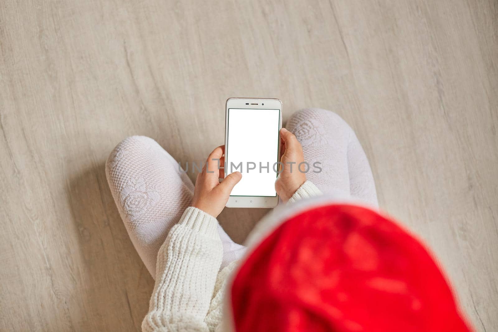 Top view of little child with smartphone with blank screen in hands hands, kid wearing santa hat, white pullover and tights, Copy space for advertisement.