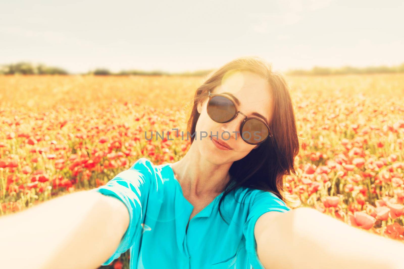 Smiling young woman in sunglasses taking selfie on background of poppies flower meadow, point of view.