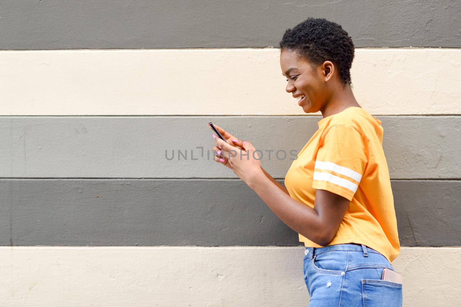 Young black woman laughing using her smart phone outdoors. Lifestyle concept. Girl with very short hair wearing casual clothes.