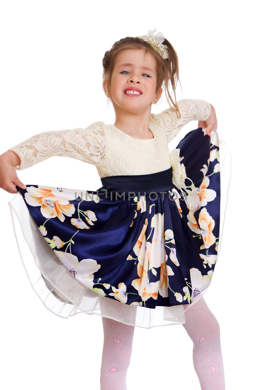 Charming girl holding the hem of her dress. Beautiful happy kid dressed nice dress and white tights standing against white background. Studio portrait of charming little girl