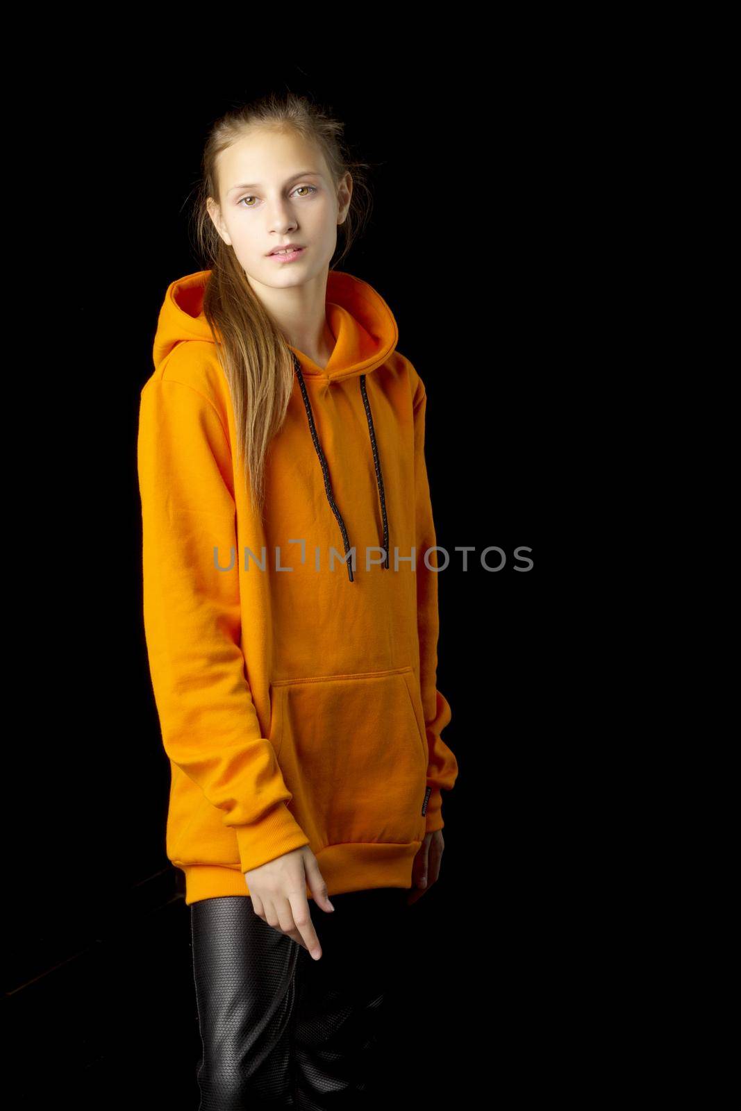 Attractive teenage girl in fashionable outfit. Three quarter length studio shot of girl with ponytail wearing oversized orange hoodie and black leather pants posing against black background