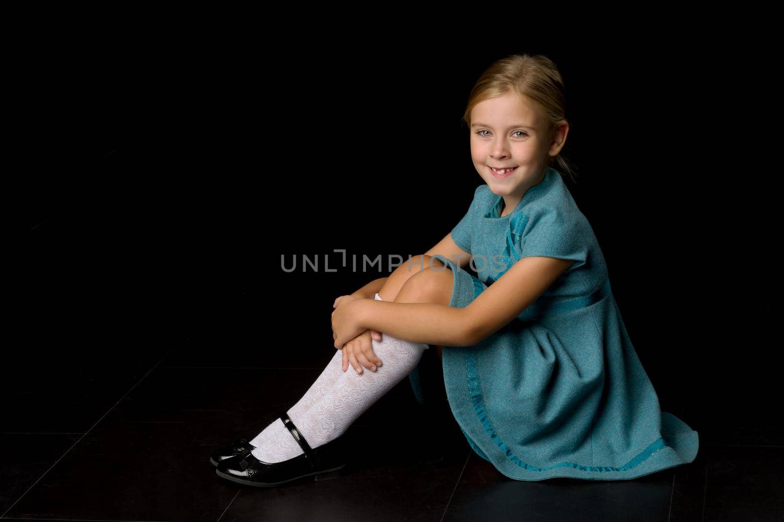 Cute girl sitting on floor hugging her knees. Adorable blonde girl wearing stylish blue dress, knee socks and black shoes sitting against black background. Portrait of charming kid posing in studio