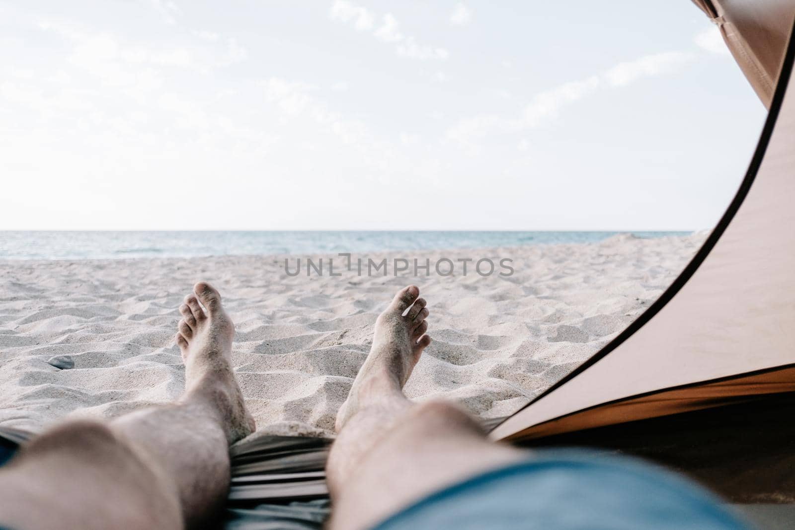 Man relaxing in a tent on sand coast in front of sea, point of view. Beach vacations.