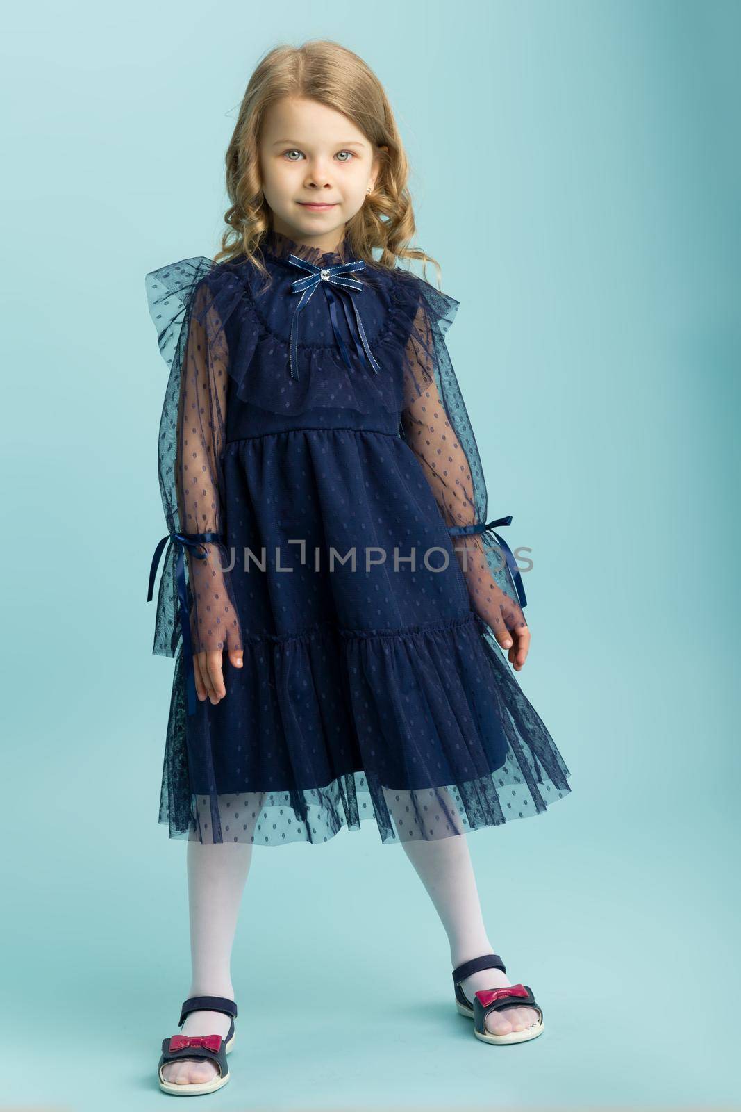 Lovely little girl posing in beautiful blue dress. Full length portrait of adorable blonde girl with curly hairstyle wearing nice lace dress standing on light blue background in studio.