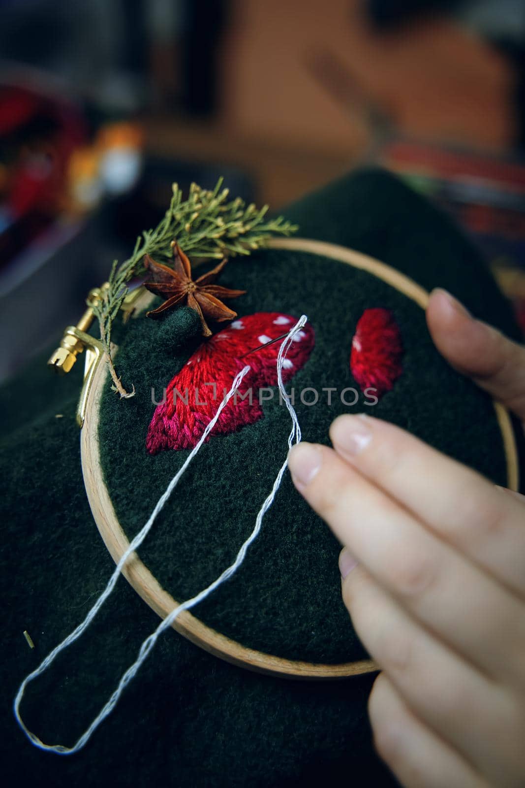 Close up of woman hands doing stitch. Process of embroidery of mushroom hat in wooden hoop on green material. Concept of needlework diy, hobby, leisure.