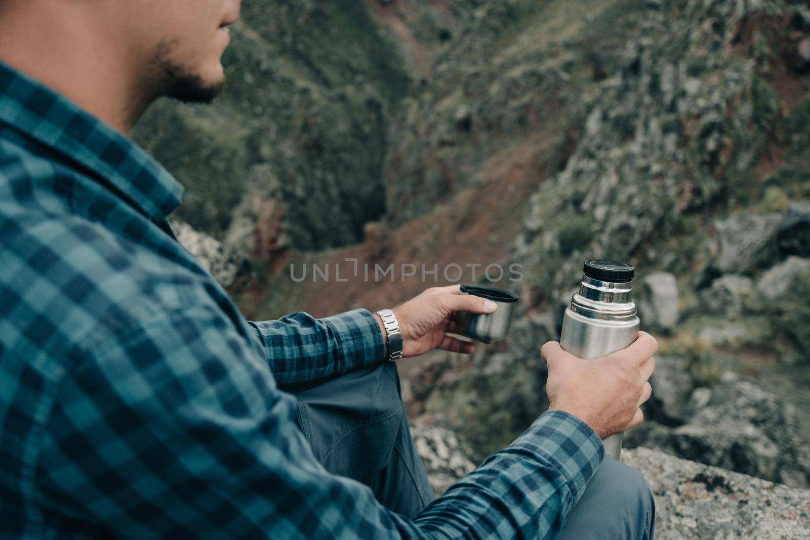 Explorer young man sitting with thermos in summer mountains outdoor, close-up.