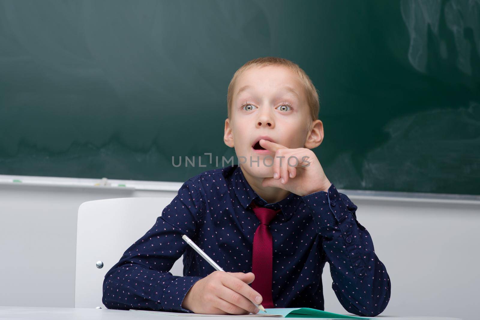 Thinking schoolboy studying at school. Boy student in blue shirt and necktie sitting at desk on background of blackboard in classroom. Back to school, education concept