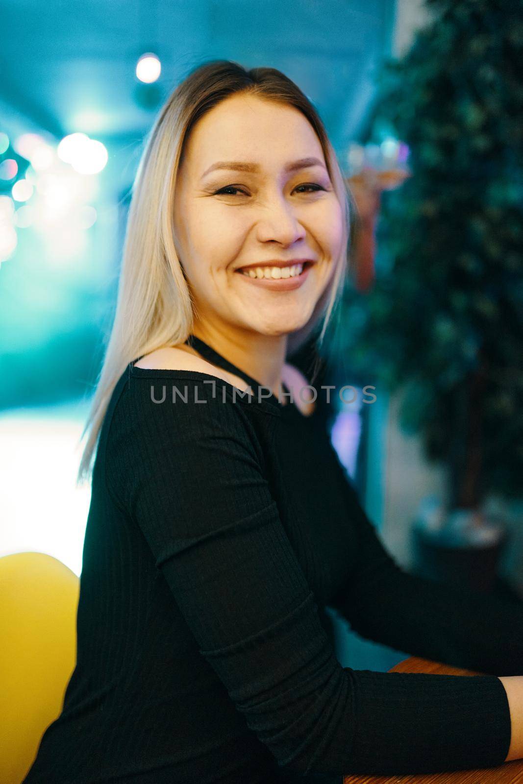 Awesome woman sits in bar and smiles. Portrait of beautiful blonde girl in black dress. Cafe interior on background. Rest and fun leisure.