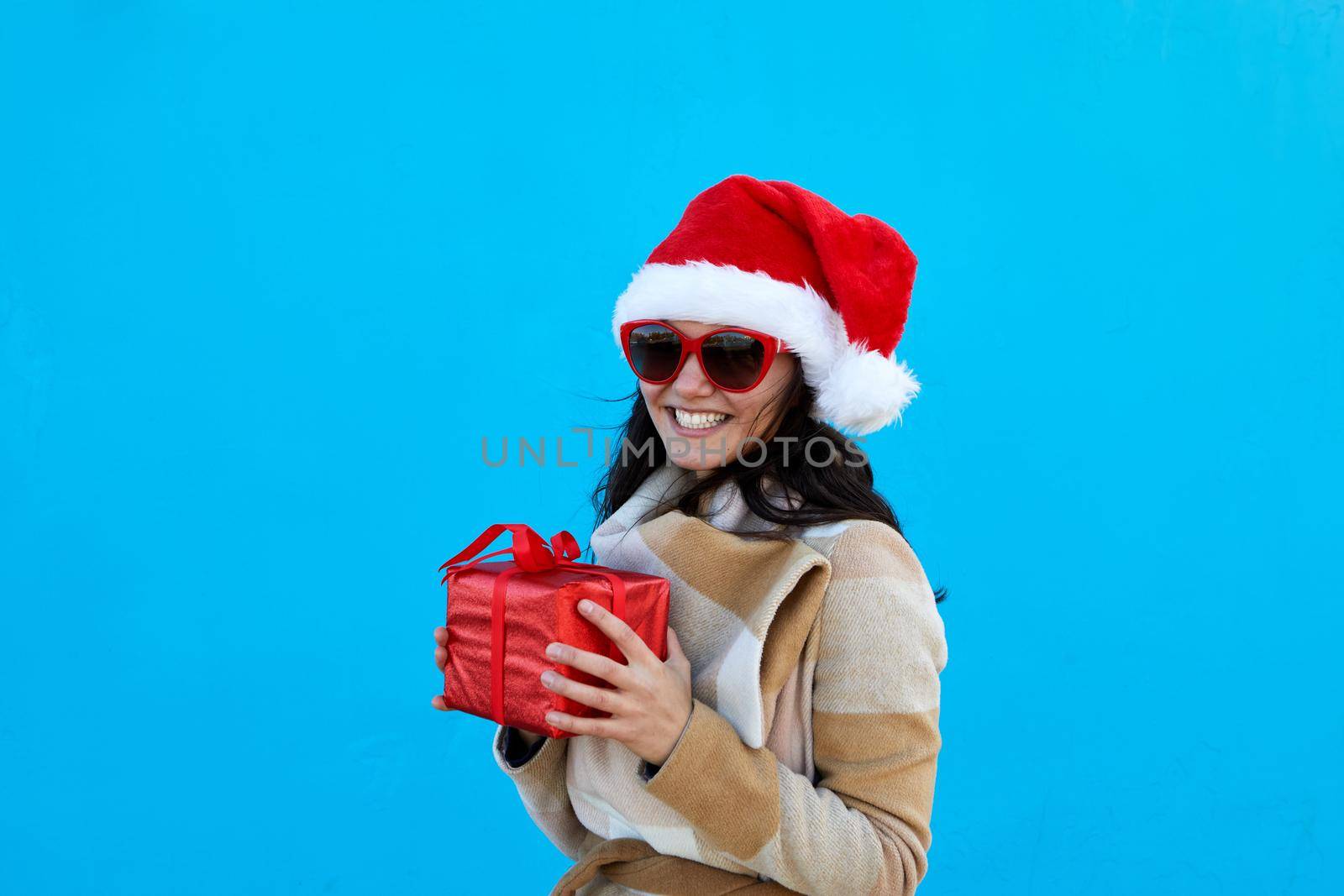 Beautiful young woman in Santa Claus hat in beige coat beret and sunglasses hold red gift over blue background