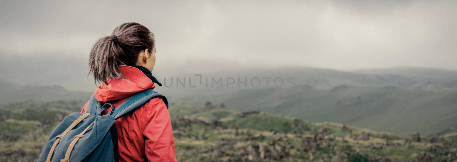 Explorer young woman with backpack walking in summer mountains in rainy weather.