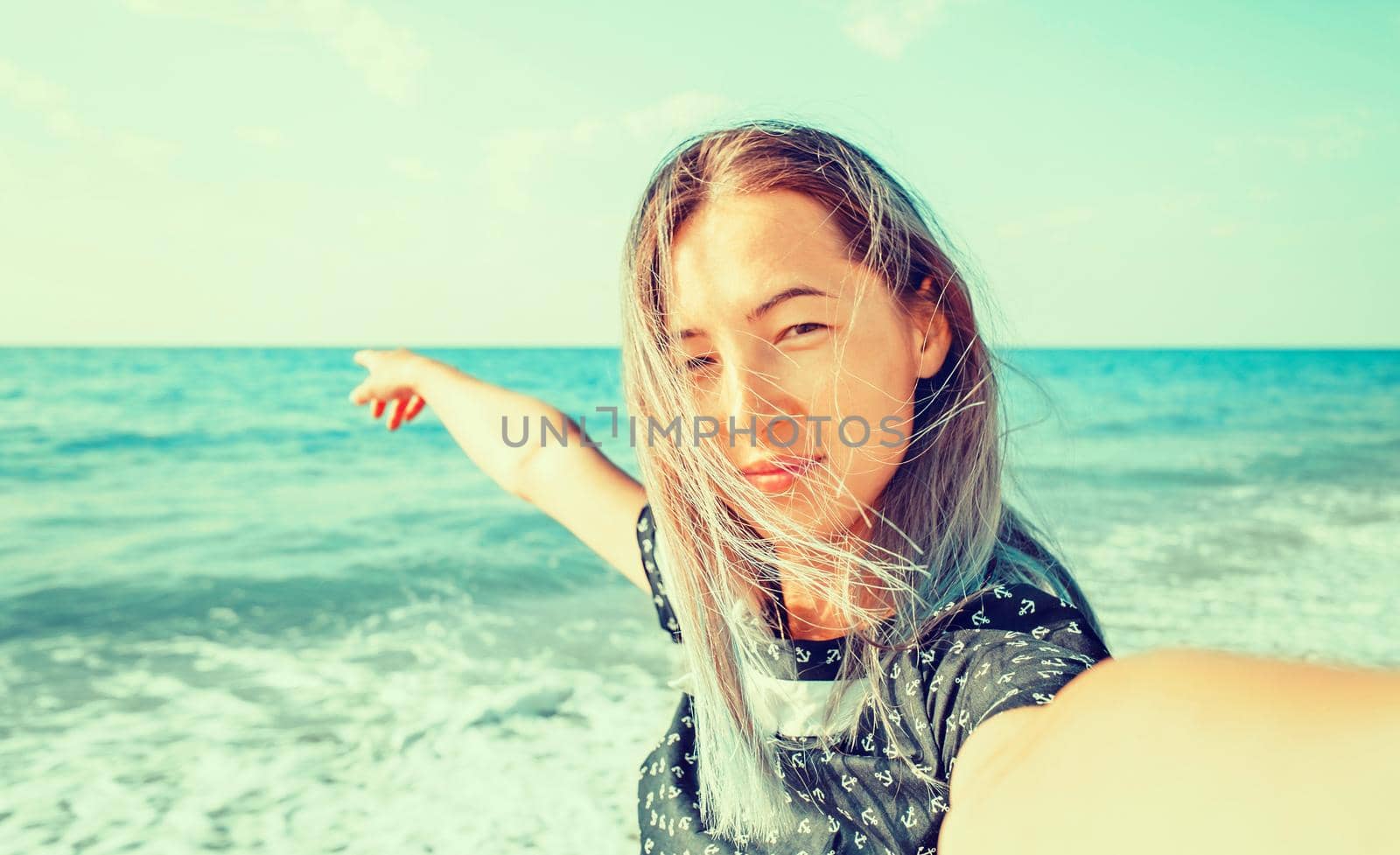 Smiling young woman looking at camera on beach and pointing at the sea, point of view shot.