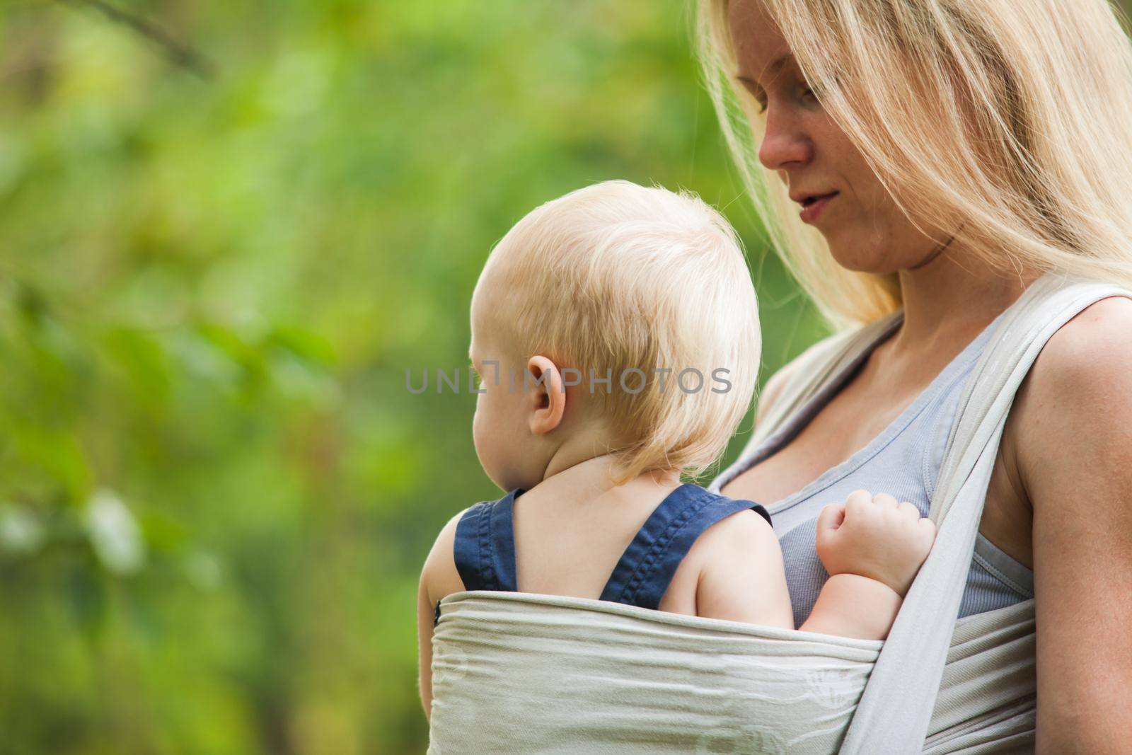 Mother is carrying her child and walking. Baby in sling outdoor.