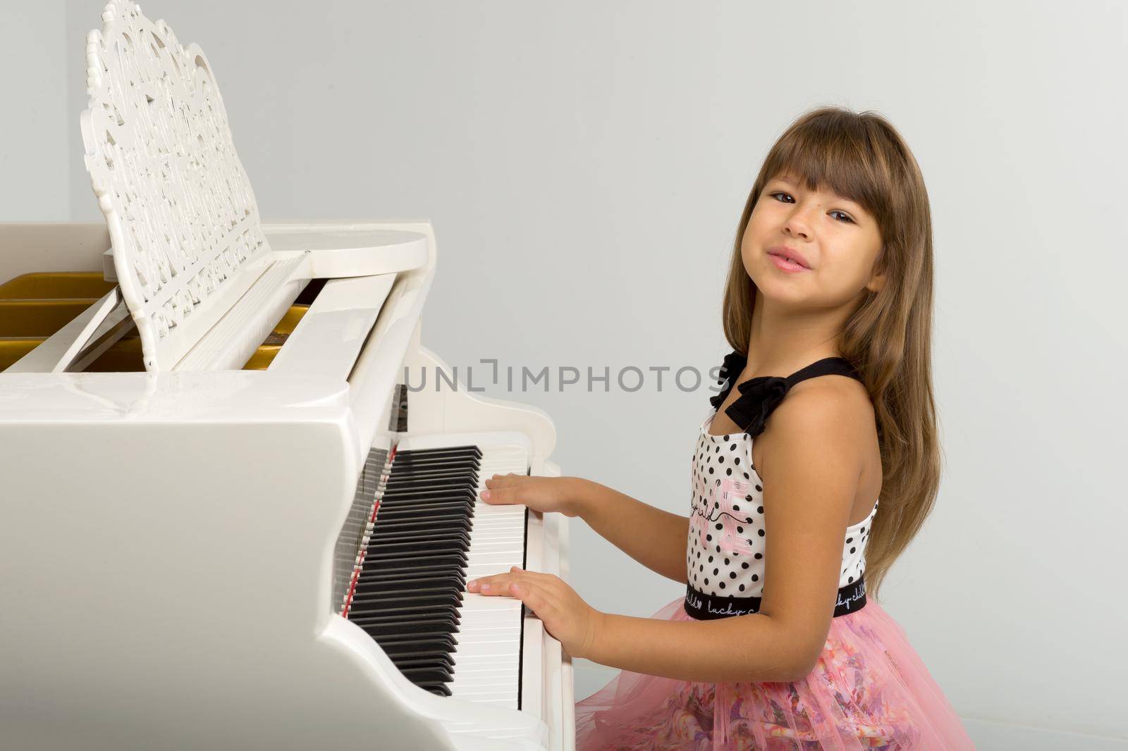Young girl posing at white grand piano. Adorable girl sitting at at musical instrument. School of arts, music lesson concept. Portrait of preteen child in nice dress looking at camera