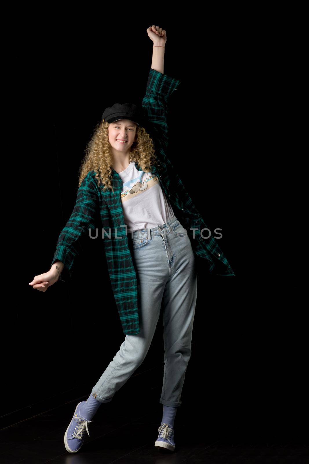 Excited girl raising her fist up in victory sign. Full length shot of happy teenager standing with her arm raised. Girl wearing plaid cotton shirt, cap and jeans posing on black background